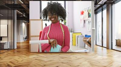 Smiling african woman tailor with glasses in a vibrant atelier, surrounded by clothing and sewing items. Wall mural