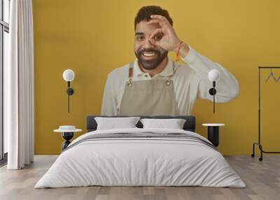 Smiling bearded man making ok gesture over eye against yellow background, wearing apron and casual attire Wall mural