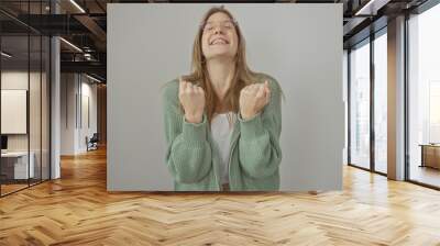 Smiling blonde woman in green sweater with clenched fists conveying excitement against a white background Wall mural