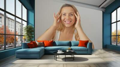 Smiling blonde woman touching face against a white isolated background, exuding happiness and confidence. Wall mural