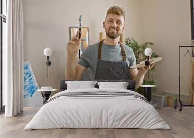 Smiling caucasian man with a beard, blue eyes, and an apron holding a brush and palette in an art studio. Wall mural