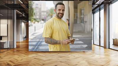 Smiling young hispanic man with a beard standing on a city street holding a smartphone, showcasing urban lifestyle. Wall mural