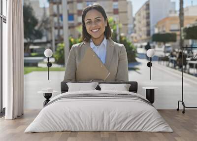 Smiling young hispanic woman in business attire holding a clipboard on a city street with buildings and trees in the background. Wall mural