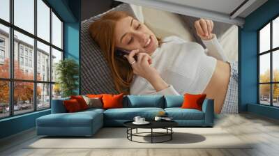 Smiling young redhead woman lying on a bed indoors, talking on the phone, wearing casual attire in a cozy bedroom. Wall mural