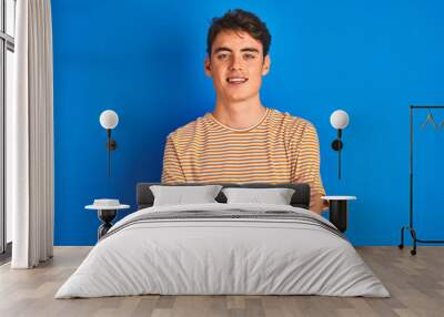 Teenager boy wearing casual t-shirt standing over blue isolated background happy face smiling with crossed arms looking at the camera. Positive person. Wall mural