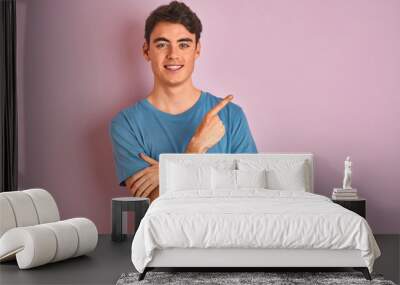 Teenager boy wearing casual t-shirt standing over blue isolated background with a big smile on face, pointing with hand and finger to the side looking at the camera. Wall mural