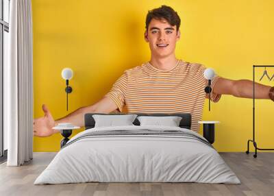 Teenager boy wearing yellow t-shirt over isolated background looking at the camera smiling with open arms for hug. Cheerful expression embracing happiness. Wall mural