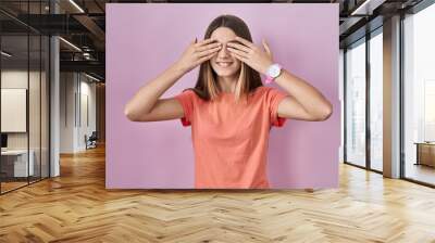 Teenager girl standing over pink background covering eyes with hands smiling cheerful and funny. blind concept. Wall mural