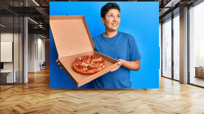 Teenager hispanic boy eating tasty pepperoni pizza smiling looking to the side and staring away thinking. Wall mural