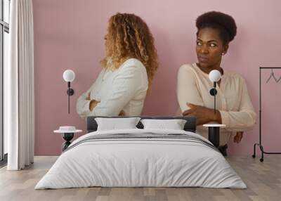 Two adult women, one african american and one latin, stand together with arms crossed, looking angry against an isolated pink background wall. Wall mural