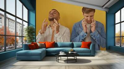 Two handsome men crossing fingers stand against a vibrant yellow wall, embodying hope and anticipation in a minimalist setting. Wall mural