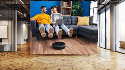 Two man couple using laptop and vacuum cleaner robot sitting on sofa at home Wall mural