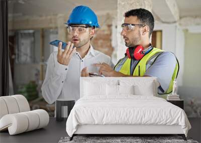 Two men builders and architect writing on document sending voice message by smartphone at construction site Wall mural