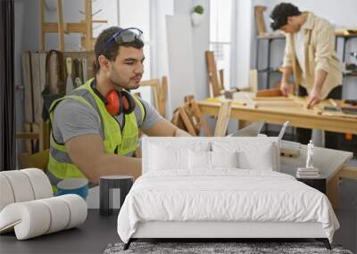 Two men working in a bright carpentry workshop, one focusing on laptop planning as his partner measures wooden planks. Wall mural