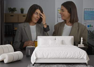 Two women in an office talking and drinking coffee, dressed in casual blazers, creating a friendly workplace environment with indoor plants in the background. Wall mural