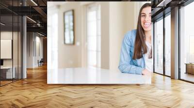 Wide angle picture of beautiful young woman sitting on white table at home sticking tongue out happy with funny expression. Emotion concept. Wall mural