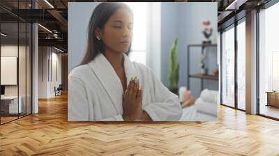 Woman meditating in a spa wearing a white robe with folded hands, showcasing beautiful nails, indoors with a serene setting, perfect for beauty and wellness themes. Wall mural