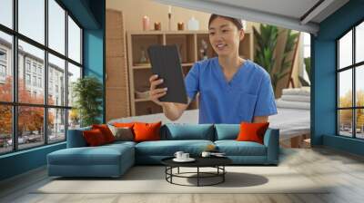 Woman smiling, beautiful, in salon spa, holding a tablet while sitting inside a wellness center with decorative plants and soft lighting, wearing blue scrubs, adult, indoors, attractive Wall mural