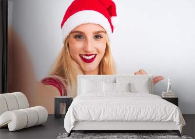 Woman wearing Christmas Santa hat make selfie by camera over isolated white background pointing and showing with thumb up to the side with happy face smiling Wall mural