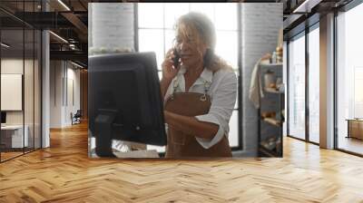 Woman working in bakery shop on phone and computer counter, shelves with assorted breads in background, wearing apron under bright indoor light Wall mural