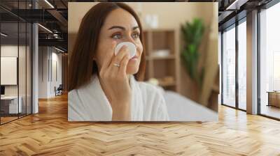 Young, woman with brunette hair in a spa room applying skincare treatment while wearing a bathrobe, surrounded by indoor wellness and beauty elements. Wall mural