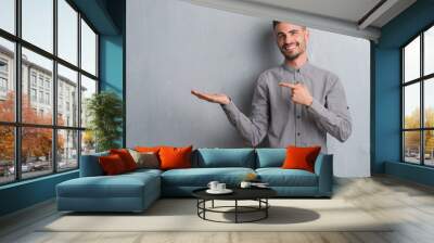 Young adult man standing over grey grunge wall amazed and smiling to the camera while presenting with hand and pointing with finger. Wall mural