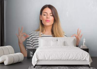 Young adult woman over grunge grey wall wearing navy t-shirt relax and smiling with eyes closed doing meditation gesture with fingers. Yoga concept. Wall mural