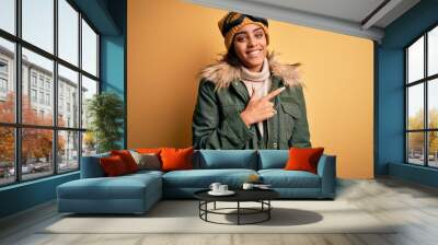 Young african american afro skier girl wearing snow sportswear and ski goggles cheerful with a smile on face pointing with hand and finger up to the side with happy and natural expression Wall mural