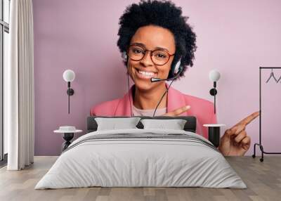 Young African American call center operator woman with curly hair using headset smiling and looking at the camera pointing with two hands and fingers to the side. Wall mural