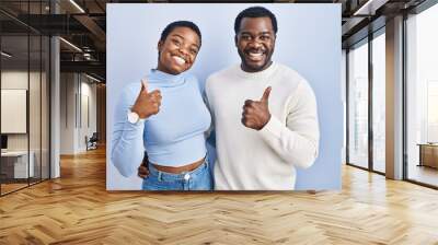 Young african american couple standing over blue background doing happy thumbs up gesture with hand. approving expression looking at the camera showing success. Wall mural