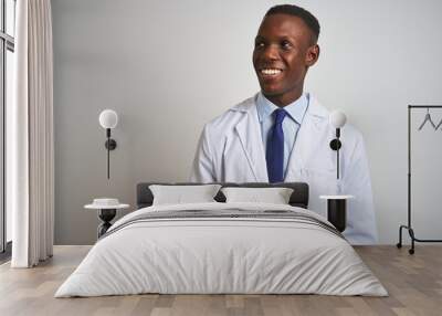 Young african american doctor man wearing coat standing over isolated white background looking away to side with smile on face, natural expression. Laughing confident. Wall mural