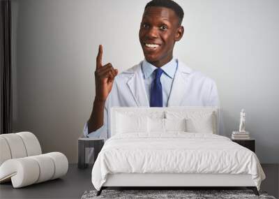 Young african american doctor man wearing coat standing over isolated white background pointing finger up with successful idea. Exited and happy. Number one. Wall mural