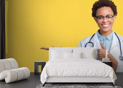 Young african american doctor woman wearing medical coat over isolated background Showing palm hand and doing ok gesture with thumbs up, smiling happy and cheerful Wall mural