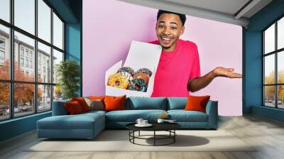 Young african american man holding box with tasty colorful doughnuts celebrating achievement with happy smile and winner expression with raised hand Wall mural