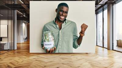 Young african american man holding jar with savings screaming proud, celebrating victory and success very excited with raised arm Wall mural