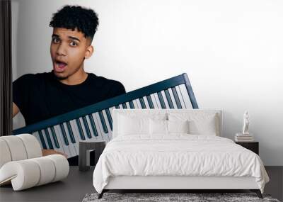 Young african american man holding piano keyboard in shock face, looking skeptical and sarcastic, surprised with open mouth Wall mural