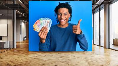 Young african american man holding swiss franc banknotes smiling happy and positive, thumb up doing excellent and approval sign Wall mural