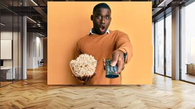 Young african american man holding television remote control eating popcorn afraid and shocked with surprise and amazed expression, fear and excited face. Wall mural