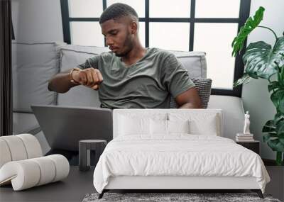 Young african american man using laptop at home sitting on the sofa checking the time on wrist watch, relaxed and confident Wall mural