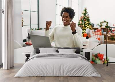 Young african american man using laptop sitting on the table by christmas tree excited for success with arms raised and eyes closed celebrating victory smiling. winner concept. Wall mural