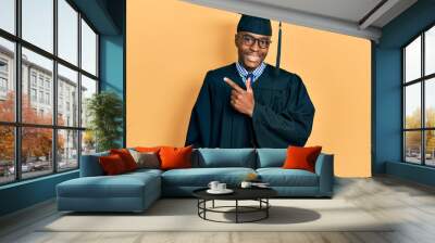 Young african american man wearing graduation cap and ceremony robe cheerful with a smile on face pointing with hand and finger up to the side with happy and natural expression Wall mural