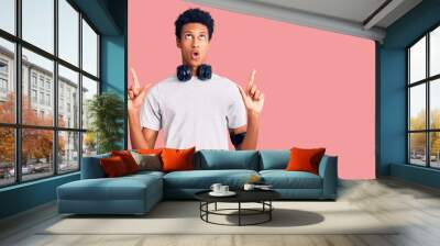 Young african american man wearing gym clothes and using headphones amazed and surprised looking up and pointing with fingers and raised arms. Wall mural