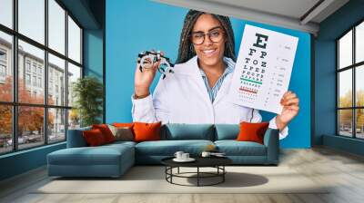 Young african american optician woman with braids holding optometry glasses and medical exam winking looking at the camera with sexy expression, cheerful and happy face. Wall mural