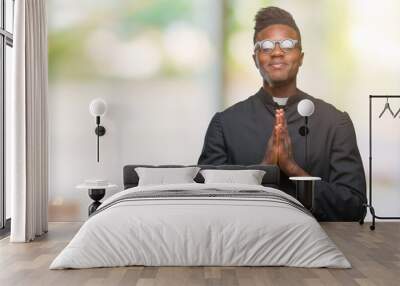 Young african american priest man over isolated background praying with hands together asking for forgiveness smiling confident. Wall mural