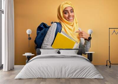 Young african american student girl wearing muslim hijab and backpack holding notebook very happy pointing with hand and finger Wall mural