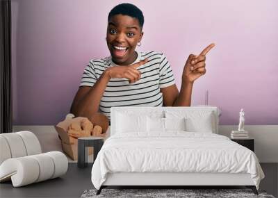 Young african american woman eating a tasty classic burger with fries and soda smiling and looking at the camera pointing with two hands and fingers to the side. Wall mural