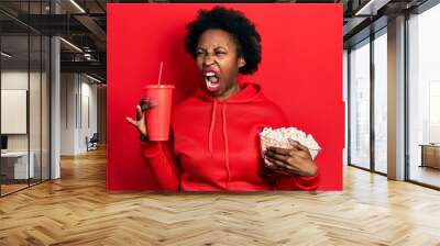 Young african american woman eating popcorn and drinking soda angry and mad screaming frustrated and furious, shouting with anger. rage and aggressive concept. Wall mural