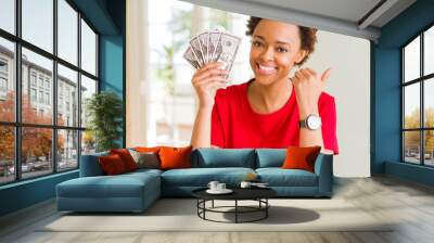 Young african american woman holding bank notes of fifty dollars pointing and showing with thumb up to the side with happy face smiling Wall mural