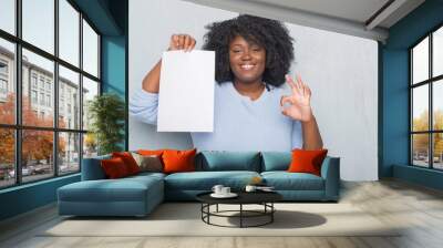 Young african american woman over grey grunge wall holding blank paper sheet doing ok sign with fingers, excellent symbol Wall mural