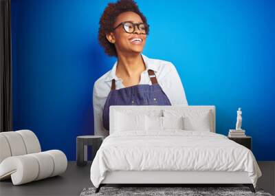 Young african american woman shop owner wearing business apron over blue background looking away to side with smile on face, natural expression. Laughing confident. Wall mural